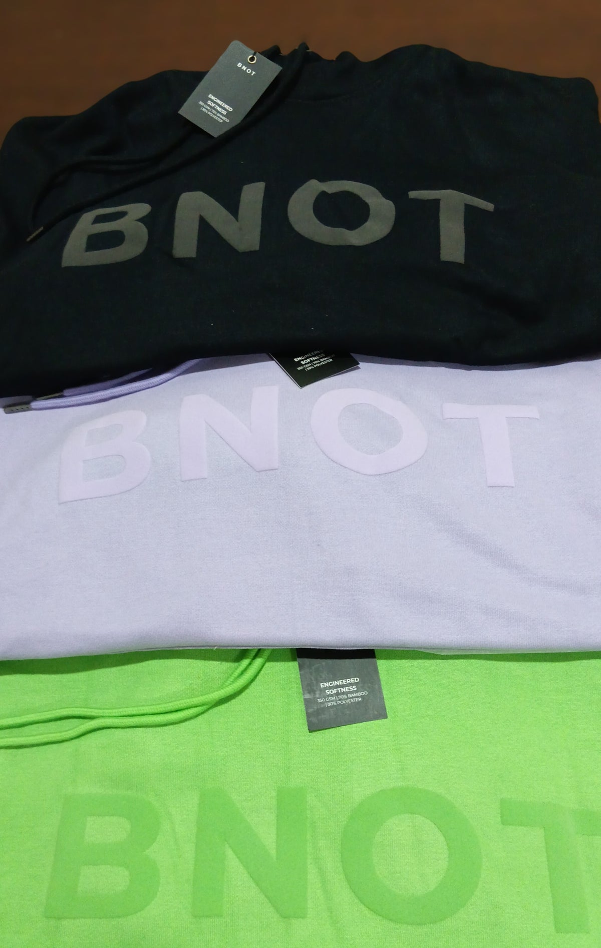 BNOT hoodies stacked in black, lavender, and green showing the 70% bamboo 30% polyester fabric tag